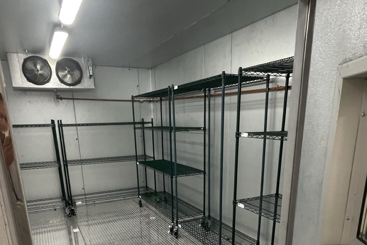 Inside a commercial walk-in freezer during a service call, showing the twin-fan ceiling evaporator, insulated panel walls, refrigerant line and diamond-plate floor